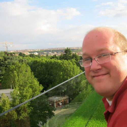 A smiling man leans over a grassy balcony rail overlooking a lush park and cityscape in bright daylight, with leafy trees and distant buildings below. He has short hair, glasses, and wears a red shirt, appearing relaxed and content in an outdoor setting.